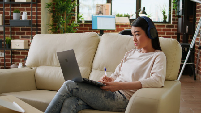 Focused working woman wearing wireless headphones taking notes while doing remote work at home sitting on sofa. Serious looking student attending online class on modern laptop.