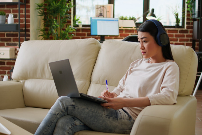 Focused working woman wearing wireless headphones taking notes while doing remote work at home sitting on sofa. Serious looking student attending online class on modern laptop.