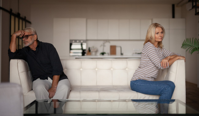 Quarrel in a mature family. Senior husband and wife are sitting on opposite sides of the sofa in the interior of the apartment. Relationship difficulties stock photo