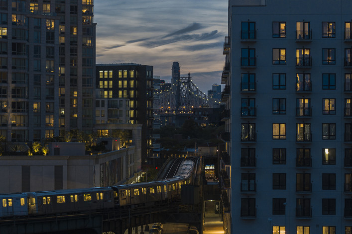 An elevated subway train riding on the line in Long Island City, Queens with Queensboro Bridge behind.