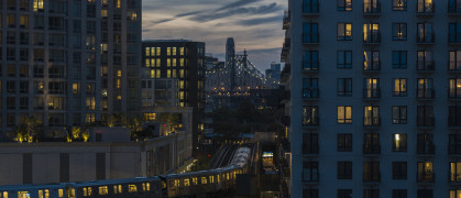 An elevated subway train riding on the line in Long Island City, Queens with Queensboro Bridge behind.